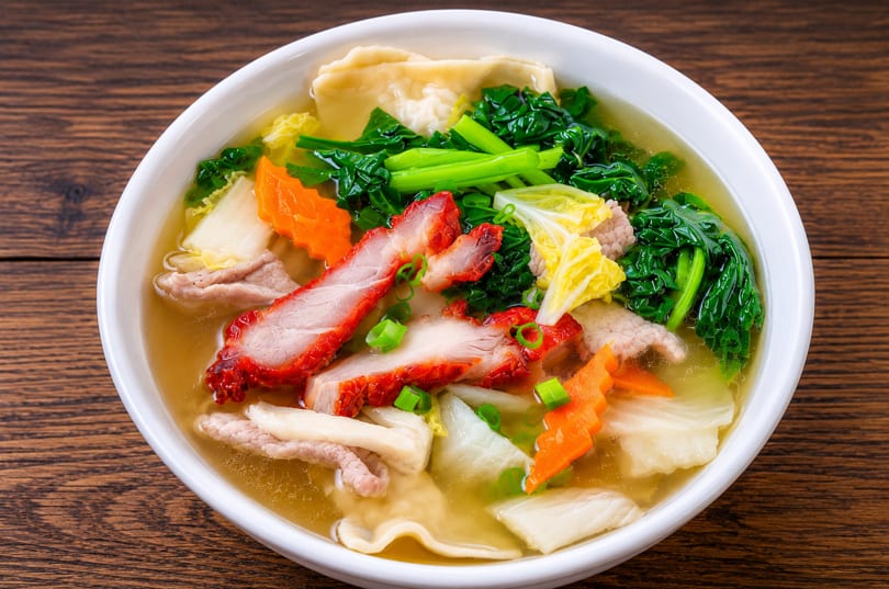 A bowl of wonton soup with sliced char siu pork, green vegetables, carrots, and noodles.